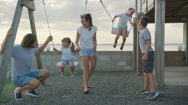 Família feliz rola crianças pequenas em um balanço ao ar livre ao pôr do sol. — Vídeo de Stock