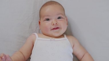 Yeni doğmuş beyaz bir bebek komik bir şekilde gülümsüyor, çocuk beşiğinin arkasında yatıyor. Neşeli ve enerjik bir çocuğun portresi.