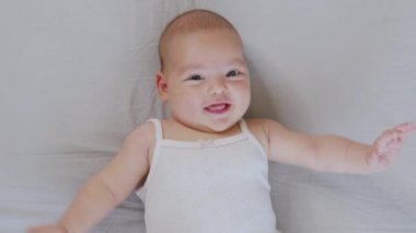 Yeni doğmuş beyaz bir bebek komik bir şekilde gülümsüyor, çocuk beşiğinin arkasında yatıyor. Neşeli ve enerjik bir çocuğun portresi.