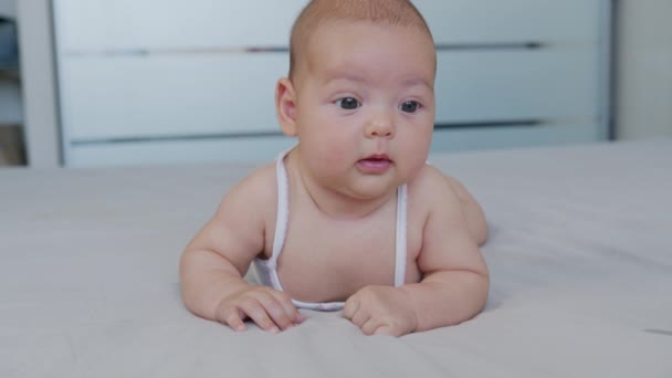 Hermoso bebé sonriente: Un bebé precioso se encuentra en la cama y sonríe a la cámara con un fondo de enfoque suave agradable. — Vídeo de stock