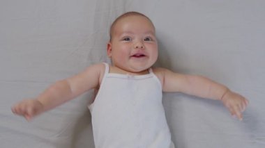 Yeni doğmuş beyaz bir bebek komik bir şekilde gülümsüyor, çocuk beşiğinin arkasında yatıyor. Neşeli ve enerjik bir çocuğun portresi.