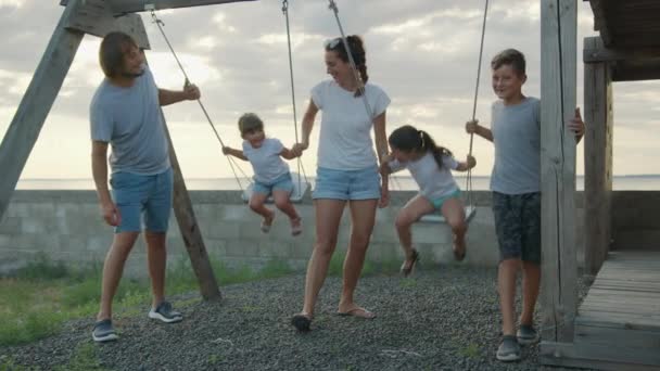 Счастливая семья катает маленьких детей на качелях на закате. — стоковое видео