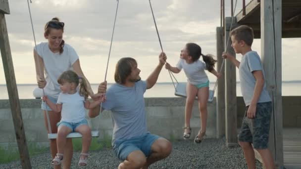 Счастливая семья катает маленьких детей на качелях на закате. — стоковое видео