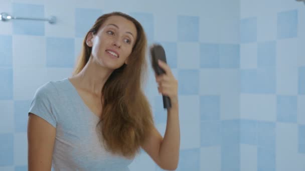 Γυναίκα κοιτάζει τον καθρέφτη, βουρτσίζει τα μαλλιά. Ερεθισμένο νεαρό κορίτσι δυσαρεστημένο με την ταλαιπωρημένη ξηρή κατάσταση των μαλλιών, απώλεια μαλλιών. — Αρχείο Βίντεο