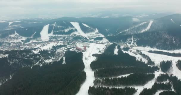 Vue aérienne des pistes de ski avec skieurs et remontées mécaniques sur la station de ski dans la forêt d'hiver contre les montagnes enneigées. Journée ensoleillée. Pistes de ski avec pins. Des collines des Carpates. Bukovel. — Video