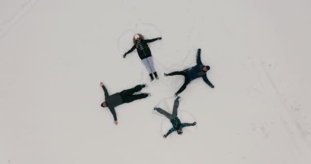 Vista aérea. Un grupo de jóvenes representa ángeles de la nieve que yacen sobre la nieve fresca en forma de cruz. cuatro ángeles de invierno felices y enérgicos con salpicaduras de nieve. — Vídeo de stock