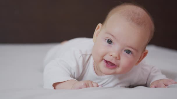 Beautiful Smiling Baby: Ένα πανέμορφο μωράκι ξαπλώνει στο κρεβάτι και χαμογελά στην κάμερα με ένα ωραίο μαλακό φόντο εστίασης. — Αρχείο Βίντεο