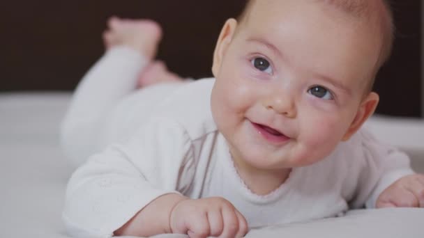 Beautiful Smiling Baby: Ένα πανέμορφο μωράκι ξαπλώνει στο κρεβάτι και χαμογελά στην κάμερα με ένα ωραίο μαλακό φόντο εστίασης. — Αρχείο Βίντεο