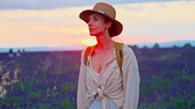 A girl tourist with a hat and a backpack walks through a lavender field during a beautiful sunset in Provence, France.