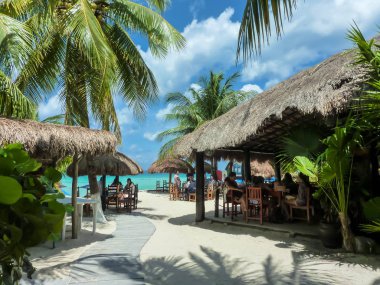 Cozumel, Meksika 24 Nisan 2019: Palancar plajında ulusal tarzda kafelerde dinlenen turistler, turkuaz denizin arka planında, parlak yeşil palmiye ağaçları ve mavi gökyüzü, Maya Meksika'daki Cozumel adası