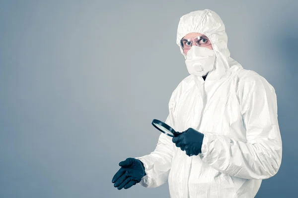 Scientist in protective clothing, glasses and respirator looks through a magnifying glass. On a gray background. Shows empty space.