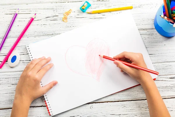 Hand of children drawing red heart with colored pencil on white paper on wooden table