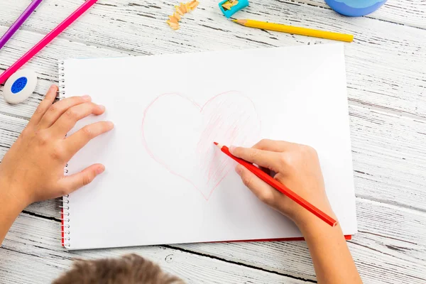 Hand of children drawing red heart with colored pencil on white paper on wooden table