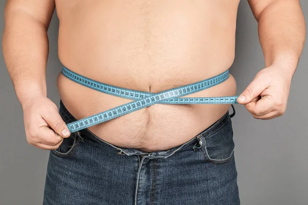 A man measures his fat belly with a measuring tape. on a gray background