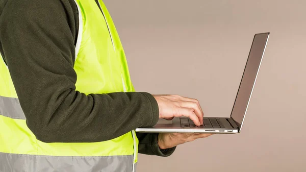 A young man, a Builder in the green safety vest, using a computer, on a gray background. Technology in industrial works