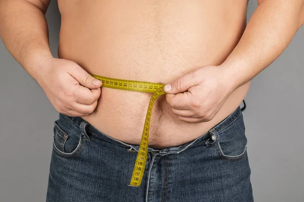 A man measures his fat belly with a measuring tape. on a gray background