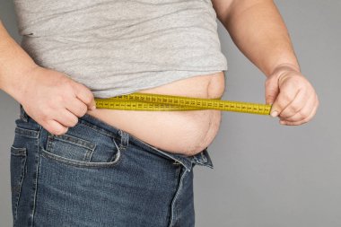 A man measures his fat belly with a measuring tape. on a gray background clipart