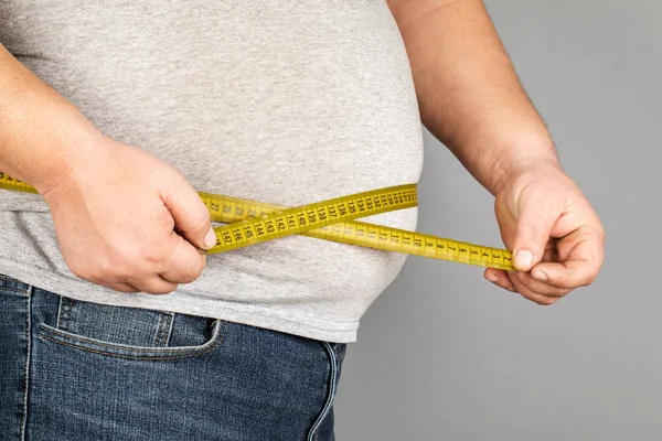 A man measures his fat belly with a measuring tape. on a gray ba — Stock Photo, Image