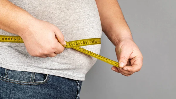 Un hombre mide su vientre gordo con una cinta métrica. en una ba gris — Foto de Stock