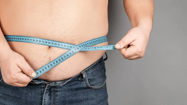 A man measures his fat belly with a measuring tape. on a gray ba — Stock Photo, Image