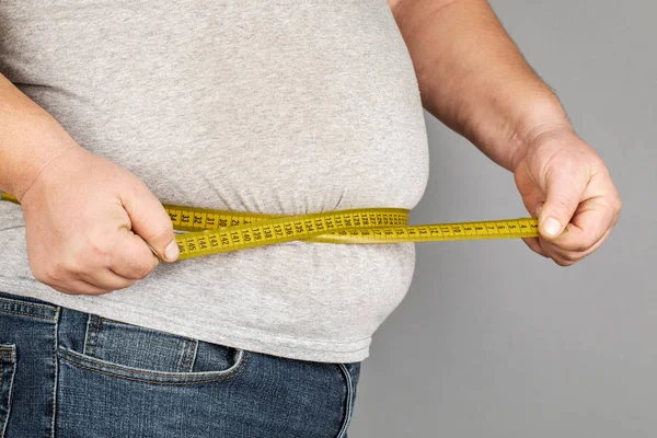 Um homem mede a barriga gorda com uma fita métrica. em uma ba cinza — Fotografia de Stock