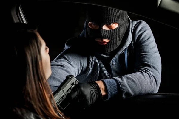 A masked robber with a gun threatens a woman in a car. robber
