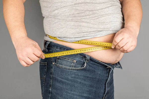 A man measures his fat belly with a measuring tape. on a gray ba