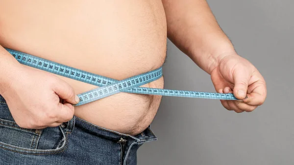 A man measures his fat belly with a measuring tape. on a gray ba