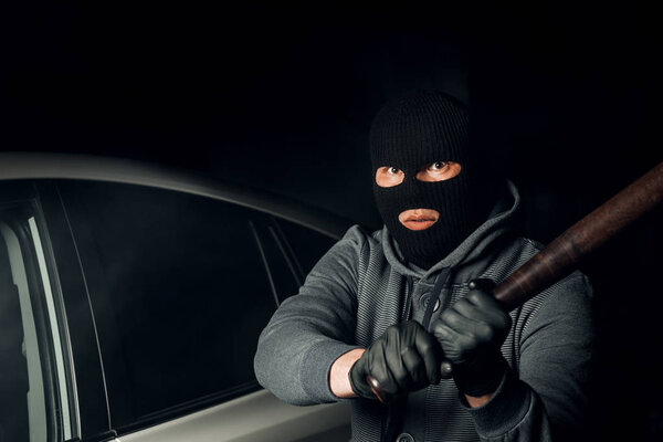 robber in a Balaclava tries to break the car glass with a baseba