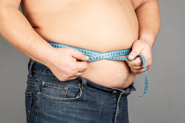 A man measures his fat belly with a measuring tape. on a gray ba