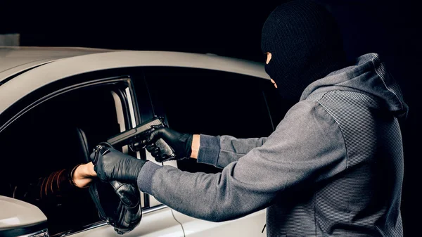 A masked robber with a gun threatens a woman in a car. robber — Stock Photo, Image