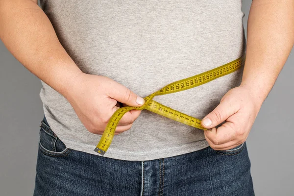 A man measures his fat belly with a measuring tape. on a gray ba