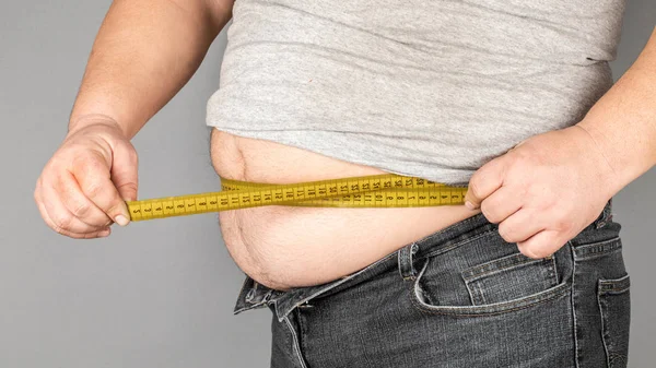 A man measures his fat belly with a measuring tape. on a gray ba — Stock Photo, Image