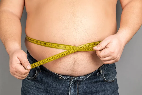 A man measures his fat belly with a measuring tape. on a gray ba