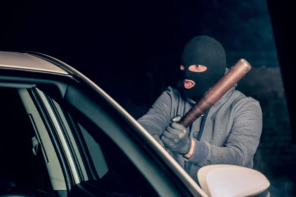 robber in a Balaclava tries to break the car glass with a baseba