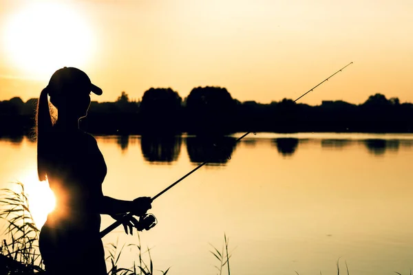 Silhuett av en kvinna vid solnedgången med ett fiskespö nära dammen — Stockfoto