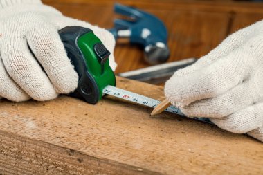 Elinde kalem ve mezura olan bir marangoz masanın üzerindeki tahta tahtayı ölçer..