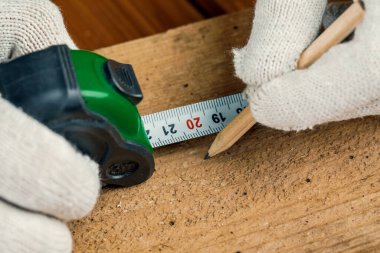 Elinde kalem ve mezura olan bir marangoz masanın üzerindeki tahta tahtayı ölçer..