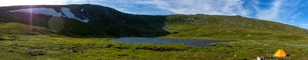 Beautiful panorama of mountain slopes with snowfields and clear mountain lake. Tent on the background of wild untouched nature of the Northern Urals. Summer trip to the mountains.