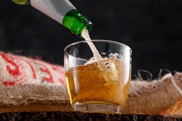 Fresh beer pouring from a bottle into a glass goblet against a textile background under burlap. Close-up.