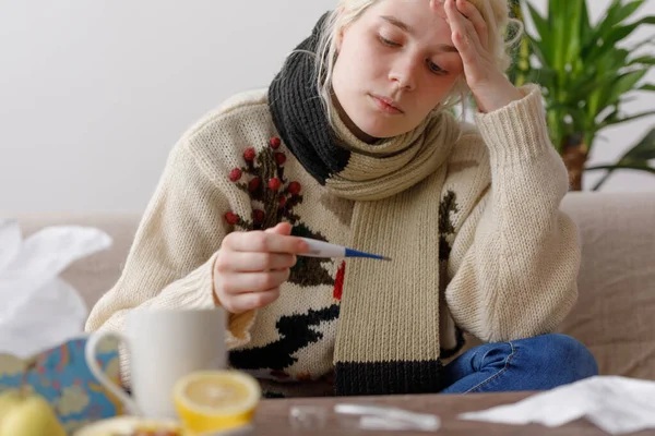 Das Mädchen im Pullover sitzt krank auf der Couch. Erkältungen und Grippe. der Patient erkältete sich, fühlte sich krank und blickte auf ein Thermometer. ungesundes Mädchen und Temperatur. — Stockfoto