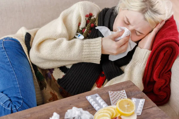 Chica en un suéter estornuda mientras está sentado en un sofá. Resfriados y gripe. El paciente se resfrió, sintiéndose enfermo y estornudando en una servilleta de papel. Una chica malsana se limpió la nariz . —  Fotos de Stock