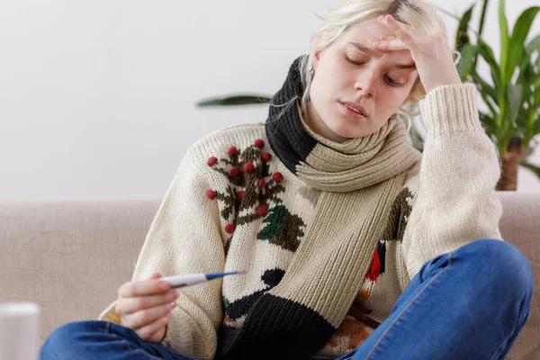 The girl in the sweater is sick sitting on the couch. Colds and flu. The patient caught a cold, feeling sick and looks at a thermometer. Unhealthy girl and temperature. — Stock Photo, Image