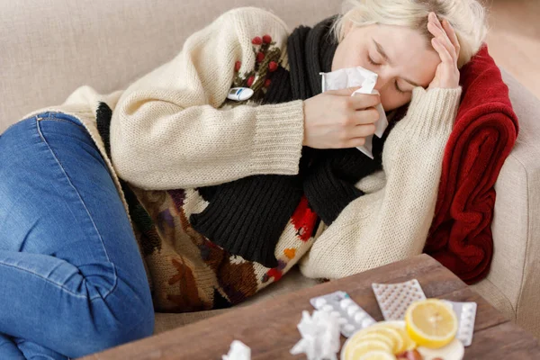 Girl in a sweater sneezes while sitting on a sofa. Colds and flu. The patient caught a cold, feeling sick and sneezing in a paper napkin. An unhealthy girl wiped her nose. — Stock Photo, Image