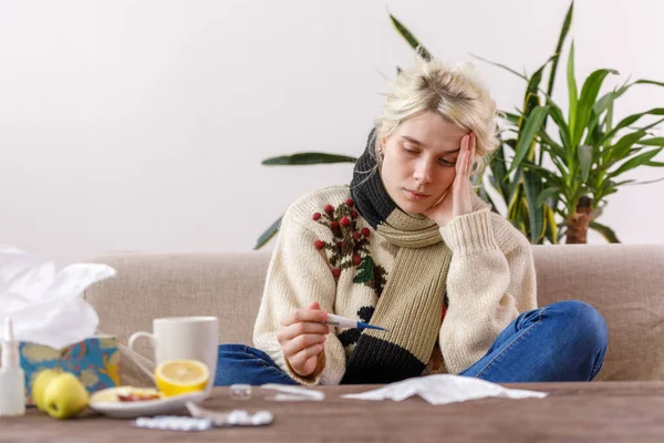 Het meisje in de trui zit ziek op de bank. verkoudheid en griep. De patiënt werd verkouden, voelde zich ziek en kijkt naar een thermometer. Ongezond meisje en temperatuur. — Stockfoto