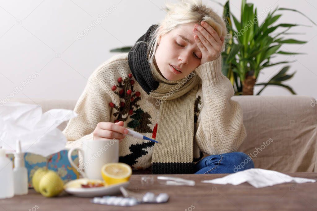 The girl in the sweater is sick sitting on the couch. Colds and flu. The patient caught a cold, feeling sick and looks at a thermometer. Unhealthy girl and temperature.