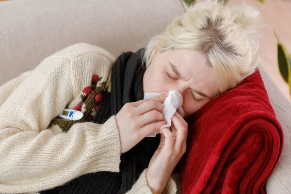 Egy lány pulóverben tüsszent, miközben a kanapén ül. Megfázás és influenza. A beteg megfázott, rosszul lett és tüsszentett egy papírszalvétában. Egy egészségtelen lány megtörölte az orrát.. — Stock Fotó
