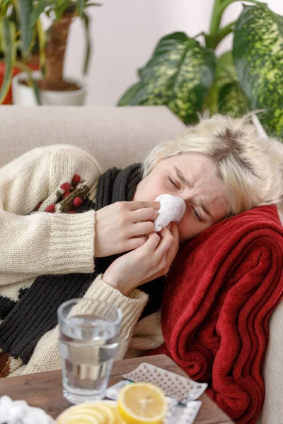 Egy lány pulóverben tüsszent, miközben a kanapén ül. Megfázás és influenza. A beteg megfázott, rosszul lett és tüsszentett egy papírszalvétában. Egy egészségtelen lány megtörölte az orrát.. — Stock Fotó