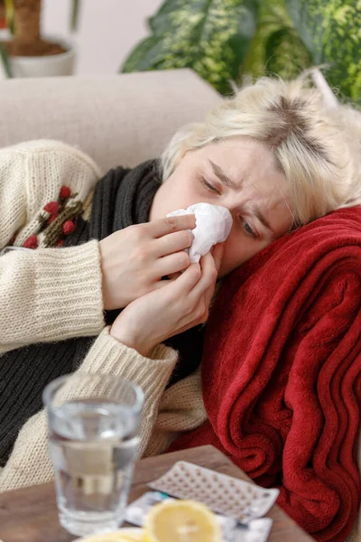 Chica en un suéter estornuda mientras está sentado en un sofá. Resfriados y gripe. El paciente se resfrió, sintiéndose enfermo y estornudando en una servilleta de papel. Una chica malsana se limpió la nariz . —  Fotos de Stock