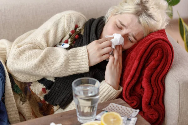 Dívka ve svetru kýchne, zatímco sedí na pohovce. nachlazení a chřipka. Pacient se nachladil, bylo mu zle a kýchal v papírovém ubrousku. Nezdravá dívka si otřela nos.. — Stock fotografie